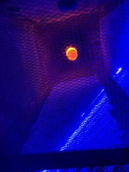 A basketball hoop is viewed from below, illuminated by vibrant blue lighting. An orange basketball is seen above the hoop, creating a striking contrast with the net and background. photo