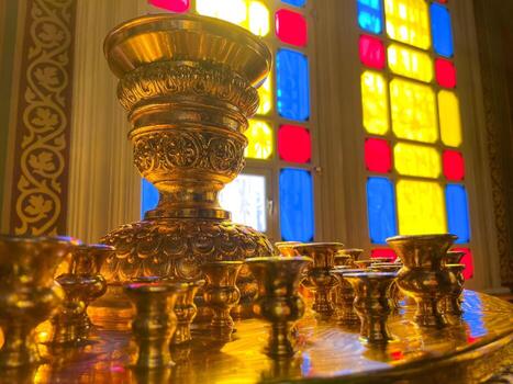 A close-up view of intricately designed brass candle holders is displayed in front of a colorful stained glass window. The background features geometric patterns in red, yellow,. photo