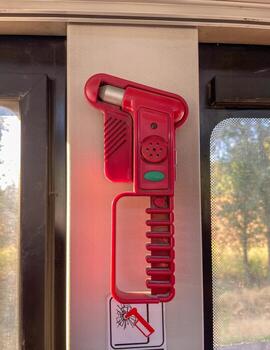 A red emergency safety hammer is mounted on a wall inside a vehicle, positioned near a window. The tool is designed for breaking glass in case of emergencies and includes a. photo