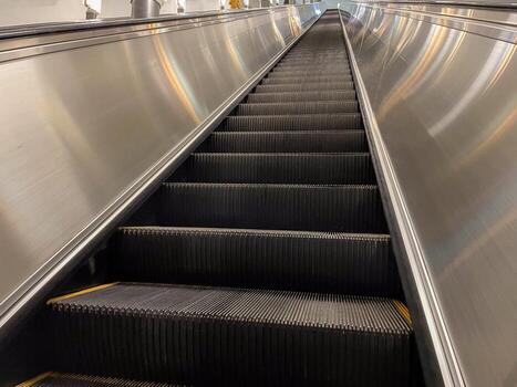 A close-up, upward perspective of an empty escalator in a well-lit indoor setting. The metallic sides and black steps create a sleek, modern appearance. photo