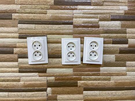 Three white double electrical outlets are mounted on a wall with a textured, multicolored tile pattern. The outlets are aligned horizontally and each has two sockets. photo