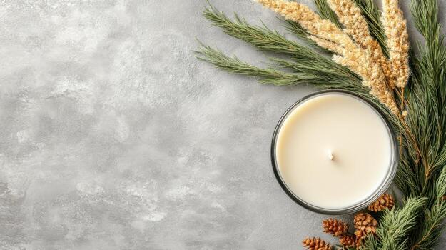 Creamy candle in clear glass with greenery and tiny pine cones on a textured gray surface providing ample copy space. photo