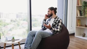 Man in his 30s speaking on phone while working remotely, using laptop and sitting at home video