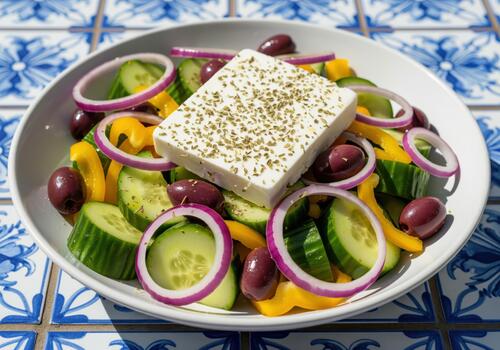 Fresh greek salad featuring a large block of feta cheese served on a patterned blue tile. photo