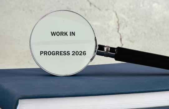 A magnifying glass rests on a stack of books, clearly showing the words work in progress 2026. This scene signifies the effort and focus on developing a future project photo