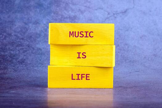Three colorful wooden blocks stand upright, each inscribed with large letters that declare music is life, set against a textured purple background. The message radiates joy photo