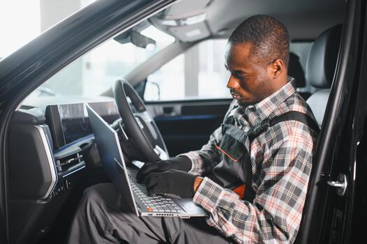 Black auto mechanic running diagnostic on, laptop while analyzing engine problems in a workshop photo