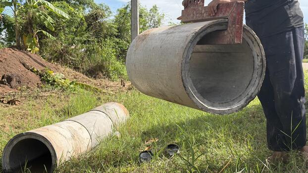 A worker lifts a heavy concrete pipe segment using machinery in an open field, preparing for the construction of an essential rural irrigation or drainage system under the midday sun. photo
