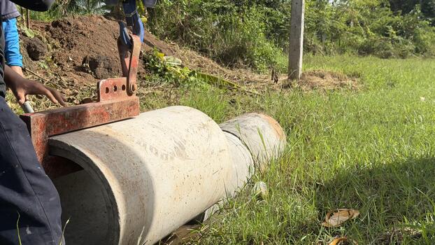 A worker guides a large concrete pipe segment being lowered by equipment, part of a construction project to establish new irrigation or drainage infrastructure in a sunny rural field. photo
