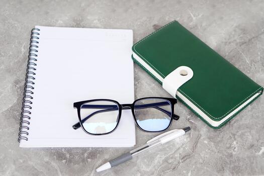 A flat lay composition featuring an open spiral notebook, a pair of eyeglasses, a green planner with a white clasp, and a pen on a gray marble surface, symbolizing organization and productivity. photo
