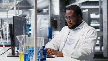 investigación instalaciones experto mecanografía en ordenador personal teclado utilizando vistoso químico soluciones en cristalería. africano americano hombre en laboratorio mirando a reactivos viales, analizando con computadora software, cámara si video