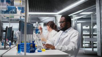 porträtt av leende laboratorium forskare använder sig av dator övervaka, bearbetning dna patient data för klinisk forskning. glad afrikansk amerikan labb arbetstagare ser på analys diagnostik på pc, kamera en video