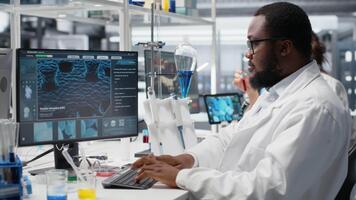Portrait of smiling laboratory scientist using computer monitor, processing DNA patient data for clinical research. Cheerful african american lab worker looking at analysis diagnostics on PC, camera B video