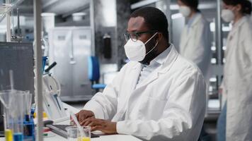 investigador vistiendo médico máscara en estéril laboratorio utilizando computadora para adn datos análisis. africano americano hombre vistiendo protector cara máscara utilizando bioinformática genómica software en ordenador personal, cámara si video