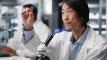 klinisk mikrobiolog granskning mikroskop glas glida i laboratorium till se celler. asiatisk labb arbetstagare checkar prov glida innan avbildning bearbeta använder sig av optisk enhet, kamera b video