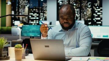 Preto geral Gerente prevendo uma tático solução e jogando com caneta, trabalhando atrasado sozinho dentro escritório. cara noivando dentro crítico pensamento, avaliando opções e soluções para dilema. Câmera b. video