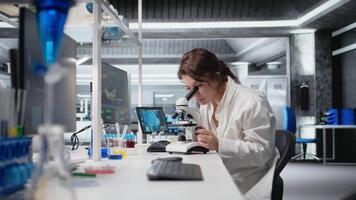 Materials scientist operating compound microscope, studying genetic disorders in laboratory . Woman studies specimen under optical device in biotechnology lab, identifying microorganisms, camera A video