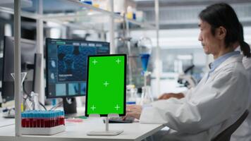 laboratorio experto a escritorio utilizando verde pantalla tableta, mirando a molecular modelo, haciendo investigación tareas. asiático hombre utilizando bioingeniería programa en Bosquejo dispositivo en médico laboratorio, cámara si video
