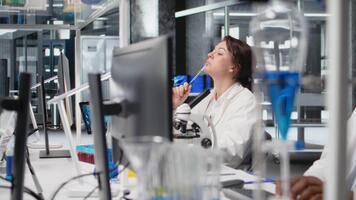 laboratorium forskare brainstorming idéer på dator skrivbord, gnuggning penna på haka, väntar för data till vara bearbetas. kvinna i labb håller på med tänkande, tolkning labb genetisk analys diagnostik, kamera en video