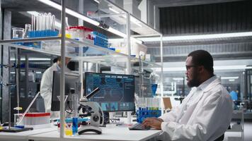 alto tecnología laboratorio técnico a escritorio mecanografía en ordenador personal teclado, mirando a molecular modelo, haciendo investigación tareas. africano americano hombre utilizando bioingeniería programa en ordenador personal en médico laboratorio, cámara un video