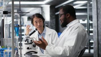 équipe de Multi-éthnique microbiologistes travail ensemble dans laboratoire avec moléculaire microscope. multiracial collègues dans recherche établissement en utilisant optique dispositif équipement à examiner médical tissu, caméra b video