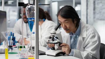 squadra capo scrive giù osservazioni per laboratorio collaboratori utilizzando microscopio per valutare cellulare strutture. asiatico laboratorio esperto prende Appunti di ingrandimento attrezzo analisi fatto di colleghi, telecamera B video