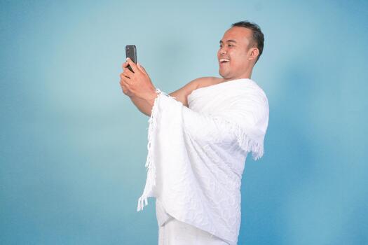 young Asian Muslim man wearing ihram, doing call pose while holding smartphone, isolated blue background. Hajj concept. islam photo