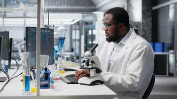 médical laboratoire technicien en utilisant scientifique microscope pour cellulaire analyse, Faire recherche Tâches. africain américain homme Faire échantillon examen à l'intérieur laboratoire avec optique dispositif engrenage, caméra b video