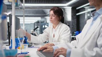 kemist använder sig av laboratorium glas fylld med indikator lösning på arbetsstation Nästa till asiatisk medarbetare. labb forskare ser på cylinder med kemisk vätska Nästa till kollega använder sig av pc, kamera en video