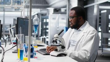 chercheur en fonctionnement composé microscope, Faire échantillon observation dans maîtrisé laboratoire environnement. africain américain homme études tissu en dessous de optique dispositif dans biotechnologie laboratoire, caméra b video