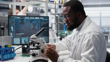 microbiologista dentro laboratório inspecionando espécime deslizar usando ótico dispositivo, analisando em pc. homem revendo citologia deslizar para clínico teste procedimento, introduzindo resultados em computador, Câmera b video