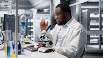 labb forskare använder sig av mikroskop till utvärdera cellulär strukturer. afrikansk amerikan man konstaterar prover genom förstoring verktyg på laboratorium bänk, framåt vetenskaplig kunskap, kamera b video