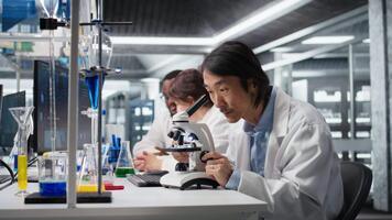 squadra capo scrive giù osservazioni per laboratorio collaboratori utilizzando microscopio per valutare cellulare strutture. asiatico laboratorio esperto prende Appunti di ingrandimento attrezzo analisi fatto di colleghi, telecamera un' video