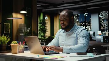 afrikansk amerikan kille svar e-postmeddelanden och dokumentera data från uppkopplad insikter för en multinationell företag, arbetssätt efter timmar i en mörk kontor. företag mål och utveckling. kamera a. video