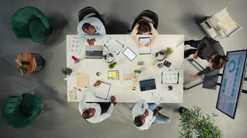 Top down view of diverse colleagues and managers participating in briefing. Business meeting focuses on negotiation, reporting and resource allocation for corporate development and success. Camera A. video