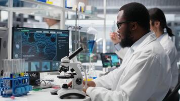 kemisk ingenjör använder sig av mikroskop till utvärdera testa prover i labb arbetsyta. forskning anläggning expert- konstaterar klinisk material genom förstoring verktyg på laboratorium bänk, kamera b video