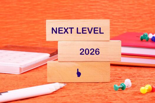 Wooden blocks stacked, displaying Next Level and 2026, with an upward-pointing finger symbol. A pencil, notebooks, and pushpins are scattered around. The background is an orange texture photo