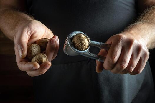 In a cozy kitchen, a person holds a handful of walnuts while using a nutcracker to split one open. The warm light highlights the earthy textures of the wood and nuts, creating a homey atmosphere photo