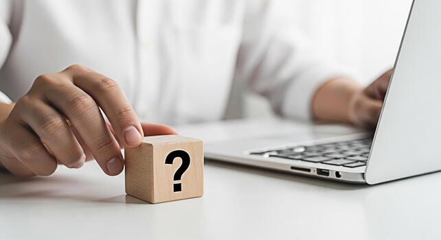 Person holding a wooden block with a question mark symbol near a laptop on a white desk representing uncertainty problemsolving and the search for answers in a modern workplace environment photo