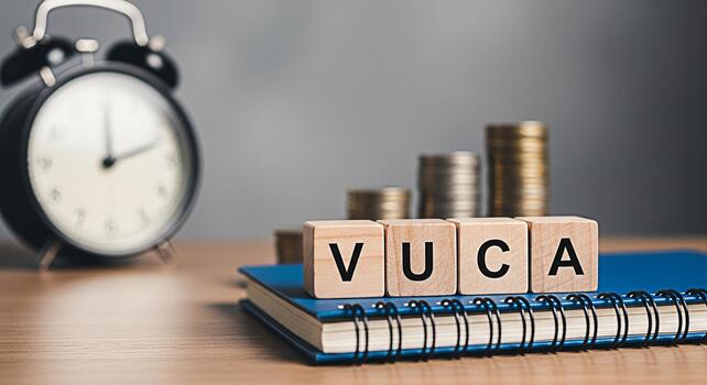 Wooden blocks spelling VUCA resting on a blue notebook with a blurred alarm clock and stacks of coins in the background representing volatility uncertainty complexity and ambiguity in business and fin photo
