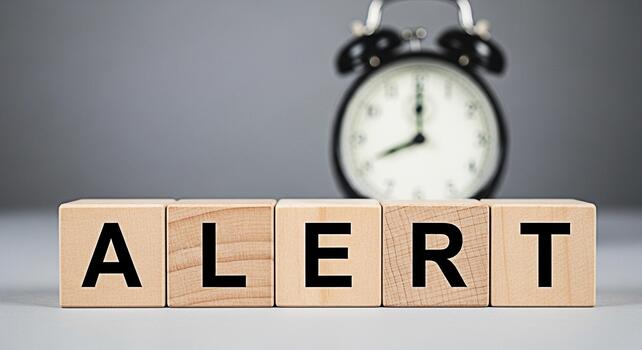 Wooden blocks spelling ALERT with a blurred alarm clock in the background symbolizing urgency and the need for immediate attention in a timesensitive situation conveying a sense of warning and importa photo