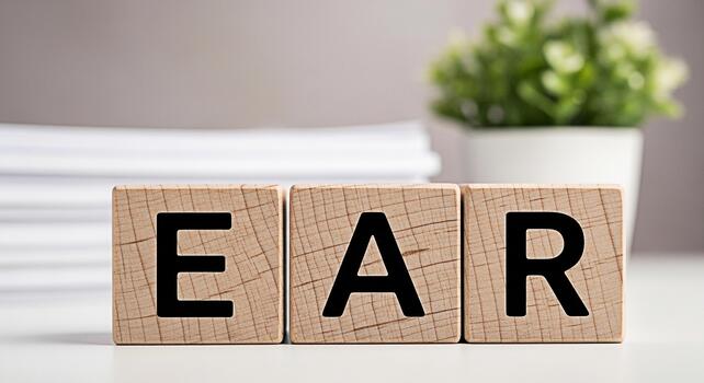 Wooden blocks spelling out EAR on a white table symbolizing hearing and listening with a plant and stack of papers in the background creating a clean and educational atmosphere in a bright room photo