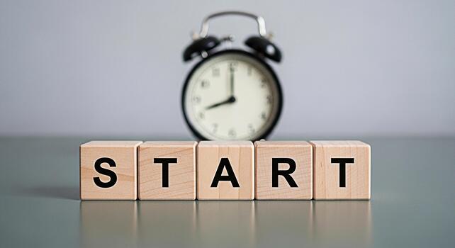 Wooden blocks spelling START with a blurred alarm clock in the background symbolizing a fresh beginning and the importance of time management in achieving goals set against a clean minimalist backdrop photo