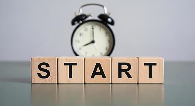 Wooden blocks spelling START with a blurred alarm clock in the background on a gray surface symbolizing a fresh beginning and the importance of time management for achieving goals photo