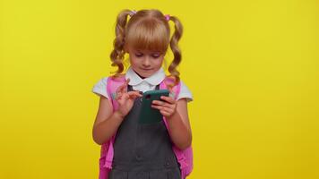 Pretty smiling blond teenage student girl dressed in school uniform using mobile phone typing new post on web, sms message, browsing, addiction of social networks on yellow studio wall background video