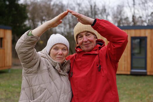 Two people stand together in a park setting, wearing warm clothing and creating a heart shape with their hands. It is a chilly day with trees in the background photo