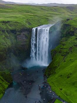 Aerial view shows Skogafoss thundering over a basalt cliff into mist. Tiny visitors stand at the base and trail. Overcast light and dark riverbed add contrast. photo