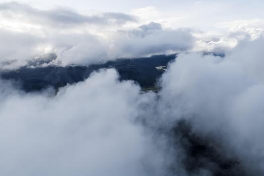 Aerial view shows thick clouds rolling over rugged highlands, a faint river visible through fog, soft diffuse light and sweeping composition suggest overcast weather. photo