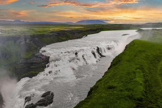 Gullfoss in southwest Iceland shows tiered cascades in a deep gorge, mist from the Hvita River, and tiny figures on a path, captured in soft golden hour light. photo