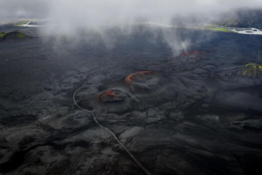 Aerial view shows steaming cinder cones, low clouds, and a gravel track on a dark lava field in Iceland, with mossy hills at edges and soft diffused light in cool weather. photo